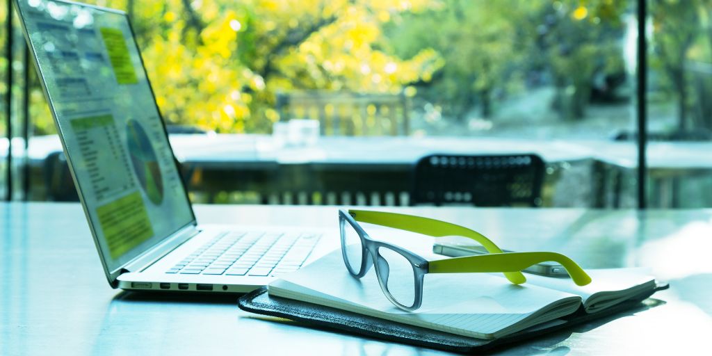 Um par de óculos apoiado em um caderno aberto ao lado de um laptop mostrando gráficos e tabelas
